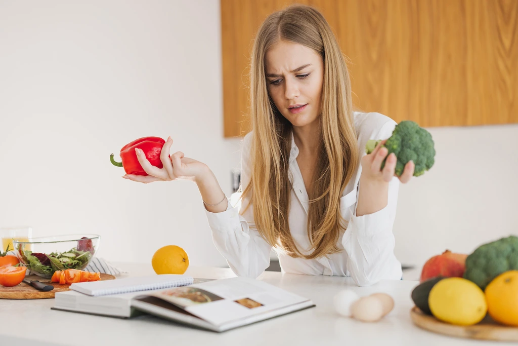 Food blogger Ava Sinclair looks comically puzzled as she holds up a red bell pepper and a head of broccoli, deciding which to use for a recipe from her cookbook.