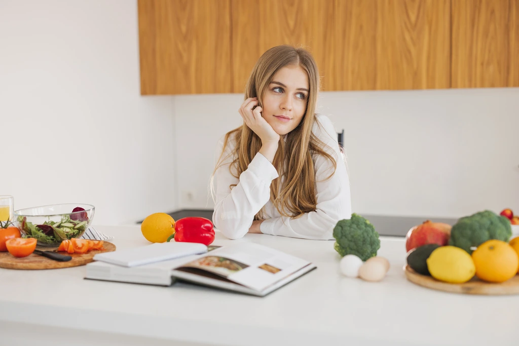 Ava Sinclair, a food blogger with blonde hair, rests her chin on her hand while brainstorming new recipe ideas at her kitchen counter, surrounded by fresh vegetables and a cookbook.