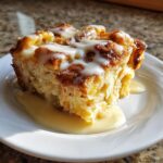 A rich, golden slice of bread pudding topped with white vanilla glaze, sitting in a pool of sauce on a white plate.