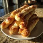 A stack of freshly baked, golden brown breadsticks sprinkled with coarse salt on a white plate.