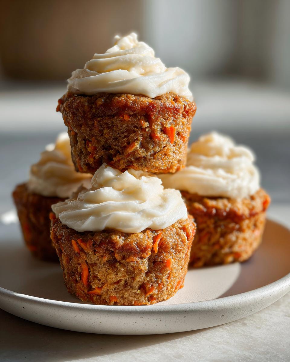 A stack of four moist carrot cake muffins topped with swirls of cream cheese frosting on a light plate.