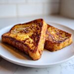 Two thick slices of perfectly golden brown french toast dusted with powdered sugar on a white plate.