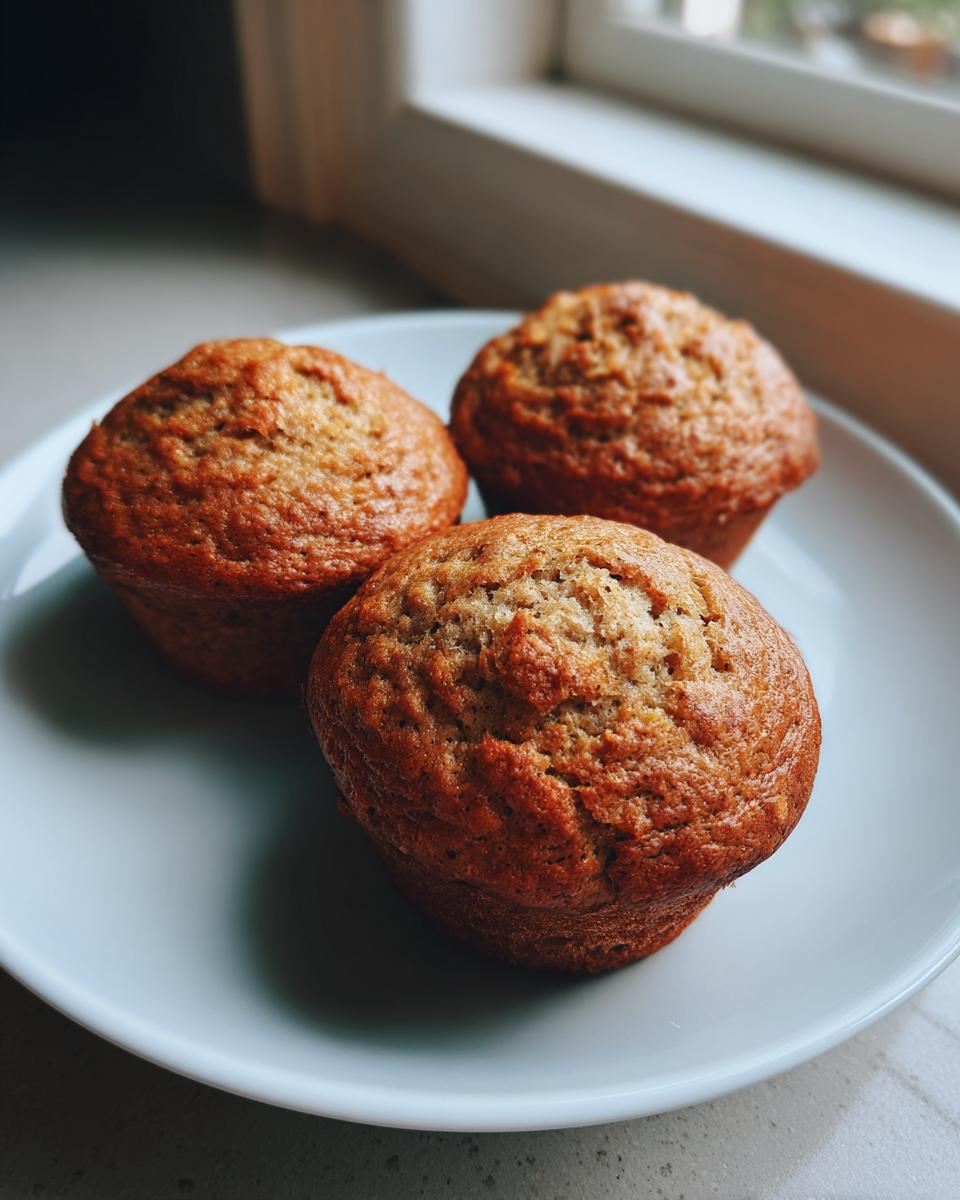 Three golden-brown banana protein muffins resting on a light blue plate near a window.