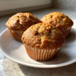 Three freshly baked banana protein muffins with golden brown tops sitting on a white plate.