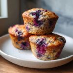 Three golden-brown blueberry protein muffins stacked on a speckled white plate.
