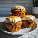 A close-up of three delicious carrot cake muffins topped with cream cheese frosting and shredded carrots.
