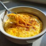 A close-up of creamy, yellow cheese grits in a white bowl, topped with shredded cheddar and spices, with a spoonful being lifted.