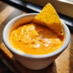 Close-up of rich, orange nacho cheese dip in a white ramekin with a single tortilla chip dipped inside.
