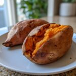 Close-up of a fluffy baked sweet potato split open on a white plate, showing bright orange flesh.