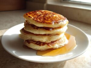 A stack of four fluffy Greek yogurt pancakes drizzled with syrup, sitting on a white plate near a window.