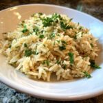 A close-up of a white bowl filled with fluffy, golden rice pilaf garnished generously with fresh chopped parsley.