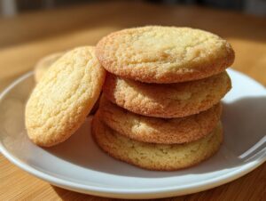 A stack of four golden, slightly textured sugar cookie rounds resting on a white plate, illuminated by sunlight.