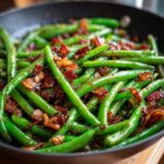 Close-up of vibrant green beans sautéed with caramelized bacon pieces in a dark skillet.