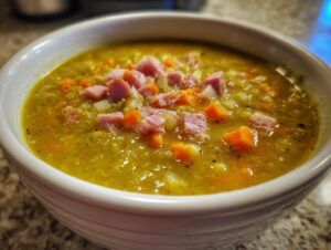 Close-up of a white bowl filled with thick, yellow-green pea soup topped with diced ham, carrots, and celery.