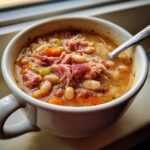 Close-up of a white mug filled with rich Ham and Bean Soup, featuring white beans, shredded ham, carrots, and celery.
