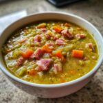 Close-up of a thick, green pea soup topped generously with diced ham and bright orange carrots.