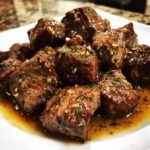 Close-up of tender crockpot steak bites coated in a rich, garlicky butter sauce on a white plate.