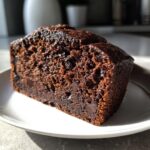 Close-up of a rich, dark slice of chocolate banana bread studded with chocolate chips on a white plate.