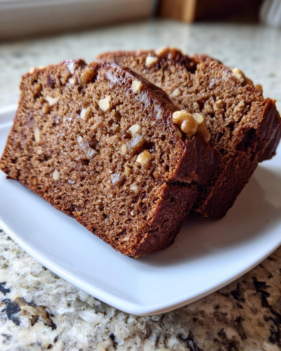 Close-up of two moist slices of dark brown date nut bread studded with walnuts, served on a white rectangular plate.