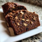 Close-up of two moist slices of dark brown date nut bread studded with walnuts on a white plate.