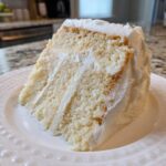 A close-up of a moist slice of vanilla cake recipe with thick white frosting layers.