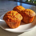 Three freshly baked morning glory muffins with visible nuts and carrots, sitting on a white plate in bright sunlight.