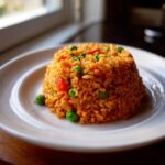 A close-up of perfectly molded, flavorful Spanish rice mixed with bright green peas and diced carrots on a white plate.