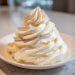 A tall, perfectly piped swirl of creamy white vanilla buttercream frosting sitting on a small white plate.
