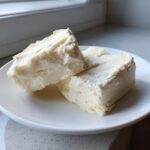 Close-up of three thick, creamy squares of white chocolate fudge resting on a white plate.