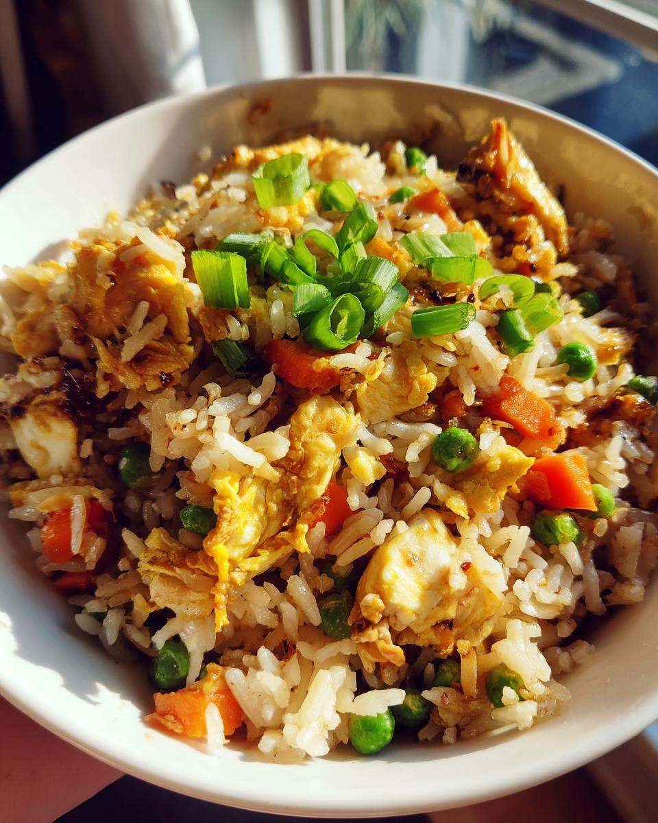 Close-up of a white bowl filled with homemade chicken fried rice, featuring scrambled egg, peas, carrots, and fresh green onions.