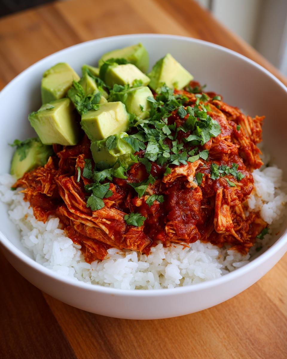 A white bowl filled with white rice, topped with saucy shredded chicken and diced avocado, ready to eat as a chicken burrito bowl.
