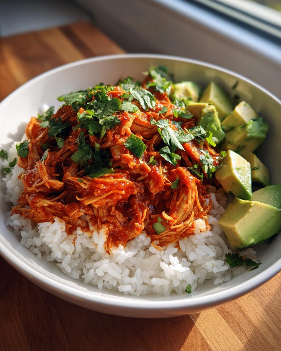 A white bowl filled with white rice, topped with saucy shredded chicken and fresh cilantro, next to chunks of avocado, making a delicious chicken burrito bowl.
