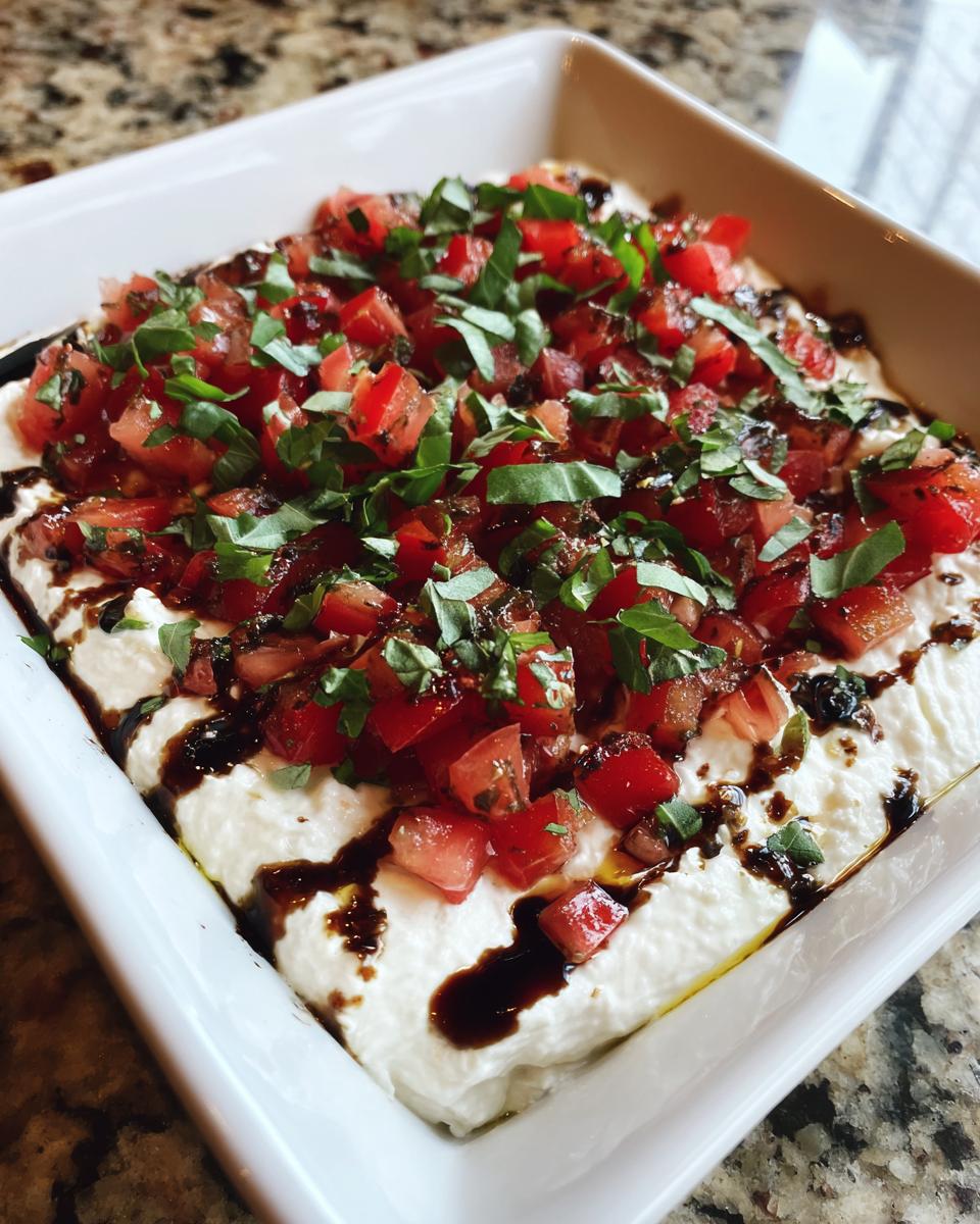 Close-up of creamy bruschetta dip topped with diced tomatoes, fresh basil, and balsamic glaze.