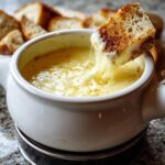 A piece of toasted bread being dipped into a white pot of hot, stringy cheese fondue.