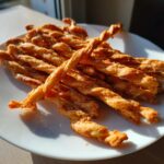 A pile of golden, crispy, twisted cheese straws resting on a bright white plate, catching the sunlight.