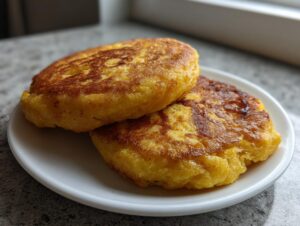 Two golden brown, perfectly seared patties of fried cornbread stacked slightly on a small white plate.