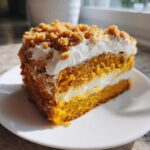 A close-up slice of moist pumpkin crunch cake layered with white frosting and topped with crunchy crumble.