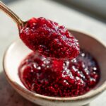 A spoonful of thick, vibrant red raspberry jam, showing visible seeds, being lifted from a small ceramic bowl.