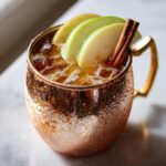 A refreshing apple cider moscow mule cocktail served over ice in a frosty copper mug, garnished with apple slices and a cinnamon stick.