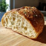 Close-up of a freshly baked artisan bread loaf, showing an open, airy crumb structure and a golden crust topped with rosemary.