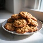 A close-up of a stack of freshly baked, golden brown banana oatmeal cookies piled high on a light gray plate.