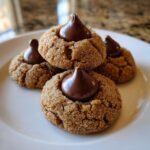 A close-up of four soft peanut butter blossom cookies rolled in sugar, each topped with a chocolate kiss.