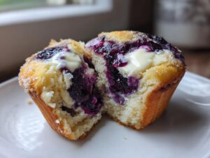 A blueberry cream cheese muffin cut in half, showing the moist interior, purple filling, and dollop of cream cheese.