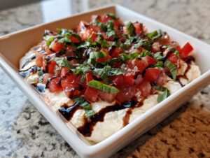 A square white dish filled with creamy bruschetta dip topped with diced tomatoes, fresh basil, and a balsamic glaze.