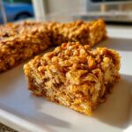 A close-up, sunlit view of a square, chewy oatmeal bars piece showing visible rolled oats texture.