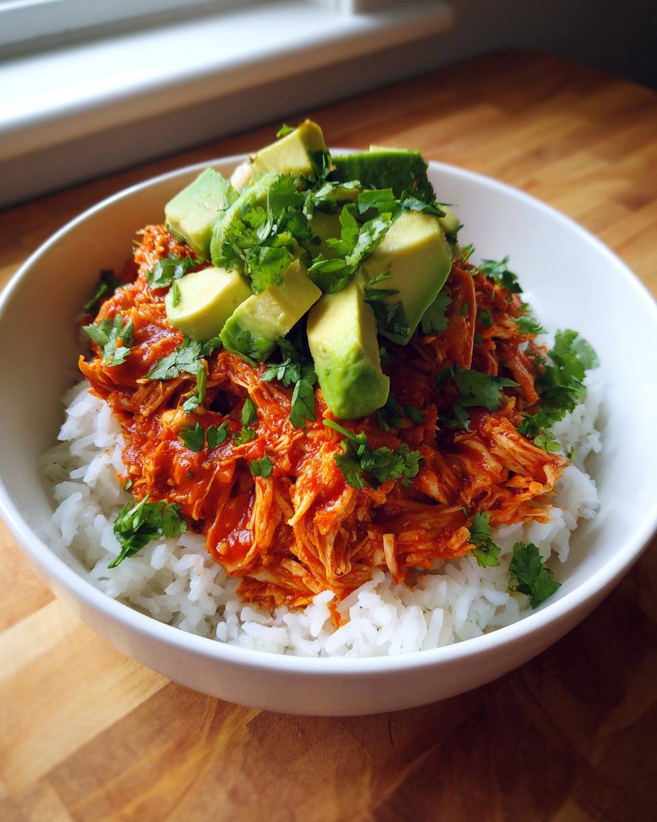 A white bowl filled with white rice, topped with shredded chicken in red sauce, diced avocado, and fresh cilantro for a chicken burrito bowl.