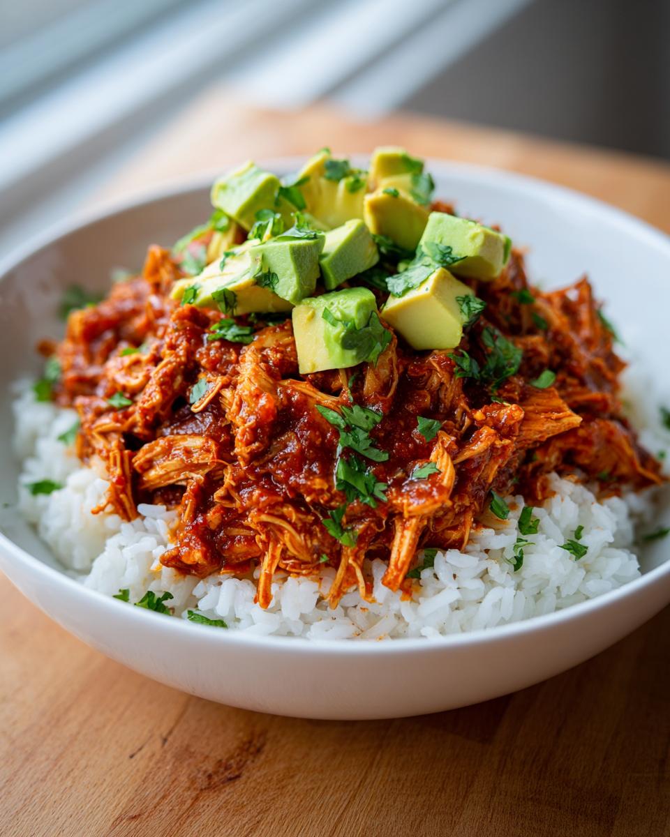 Close-up of a white bowl filled with white rice, topped with saucy shredded chicken and diced avocado for a chicken burrito bowl.