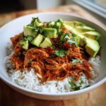 A close-up of a white bowl filled with white rice, topped with saucy shredded chicken for a chicken burrito bowl, garnished with avocado slices and cilantro.