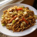A generous serving of homemade chicken chow mein recipe with noodles, chicken pieces, carrots, and cabbage on a white plate.