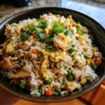 Close-up of a dark bowl filled with homemade chicken fried rice, featuring scrambled egg, peas, carrots, and green onion garnish.
